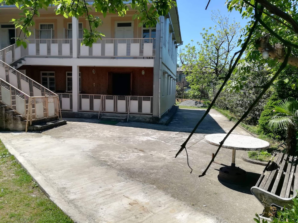 A spacious outdoor area is featured, with a concrete patio leading to a round table and bench seating. The nearby building showcases a traditional design with multiple balconies, and greenery is present in the background, contributing to a serene environment.