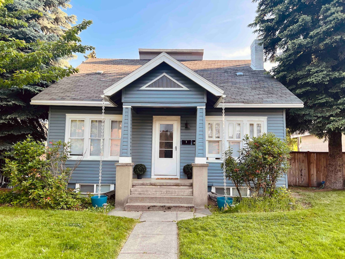 A charming blue-shingled home features a welcoming front porch with two potted plants on either side. The well-manicured lawn is framed by tall, leafy trees, adding to the inviting exterior. A pathway leads up to the front door, highlighting the home's character.