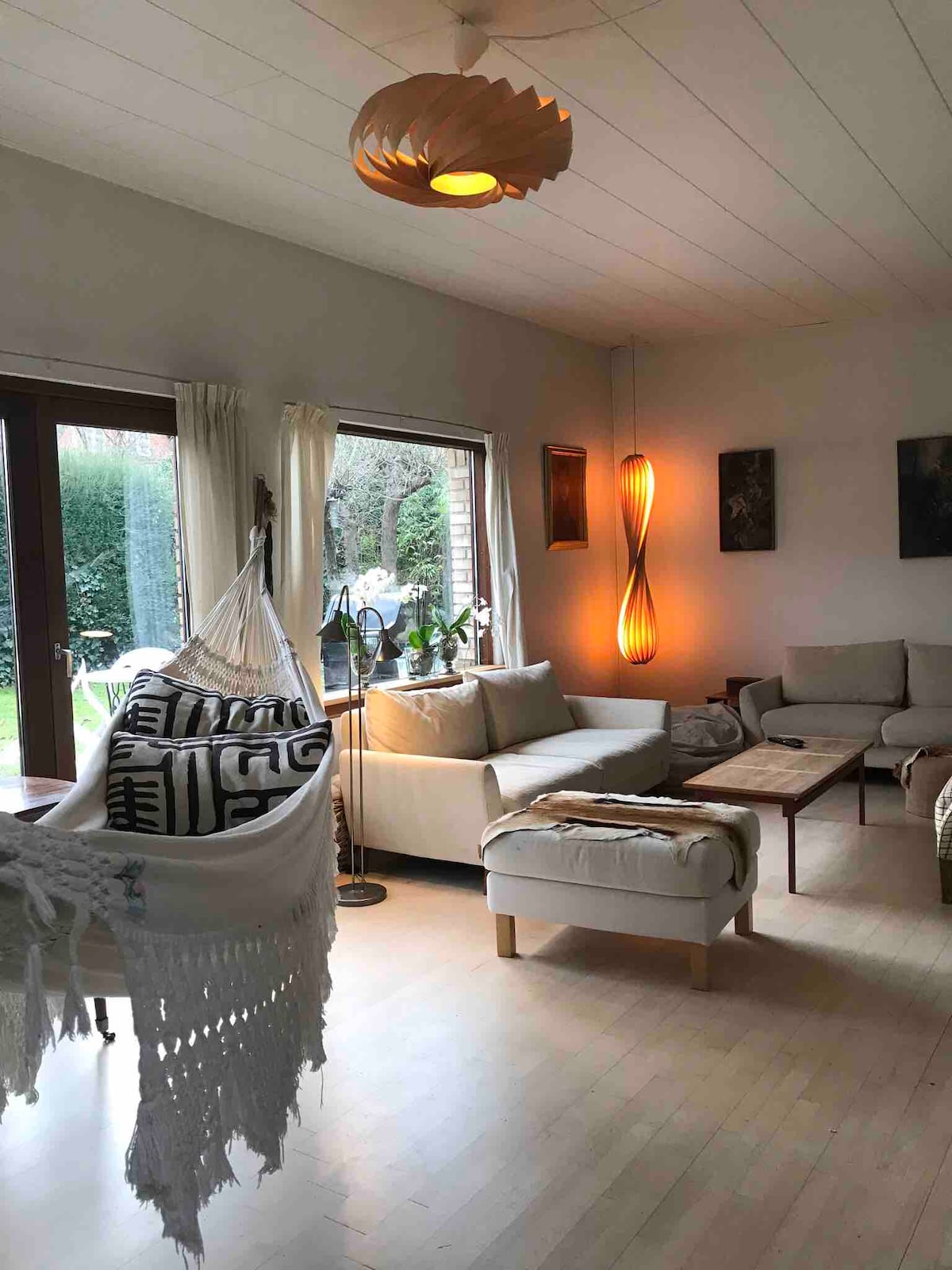 A bright living area features a comfortable seating arrangement with a mix of sofas and chairs. A stylish hammock is positioned nearby, while a warm light fixture hangs above. Large windows provide views of the garden, enhancing the space's openness.