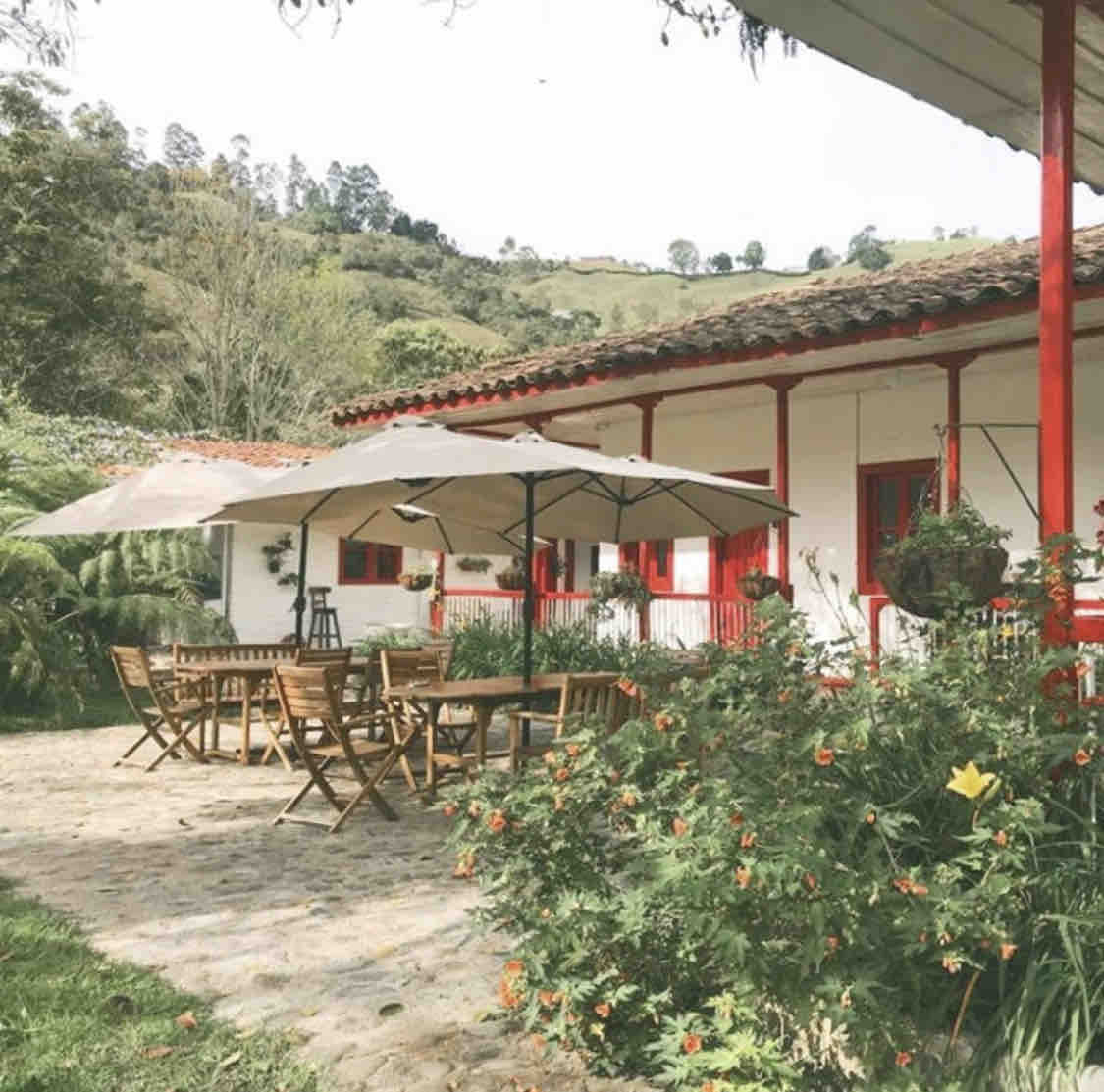A spacious outdoor area is framed by vibrant greenery and features a large table surrounded by wooden chairs. An umbrella provides shade, and the red accents of the house contrast beautifully with the white walls, enhancing the serene landscape in the background.