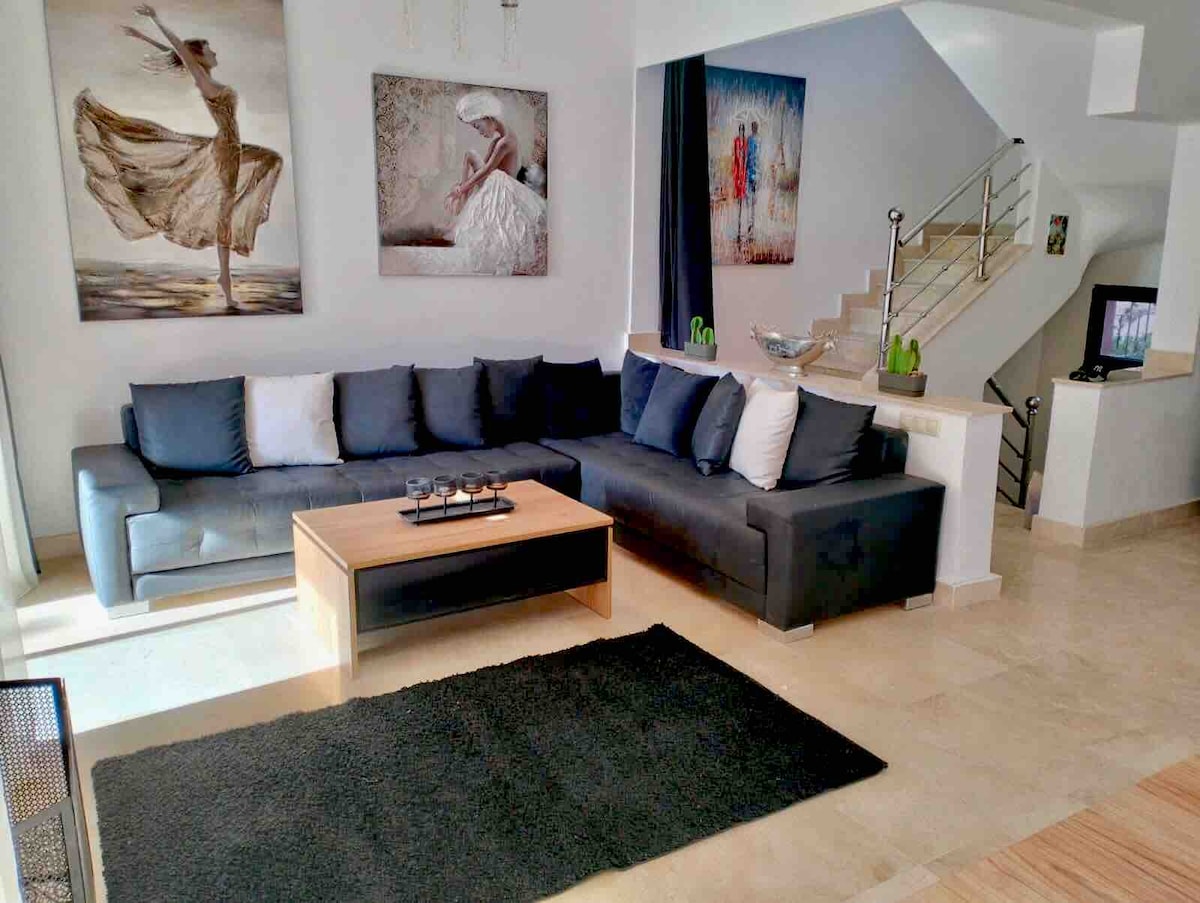 A spacious living area is presented with contemporary furnishings, featuring a dark sectional sofa with light accent pillows. A wooden coffee table sits atop a textured black rug. Colorful wall art enhances the ambiance, while natural light floods the room from nearby windows.