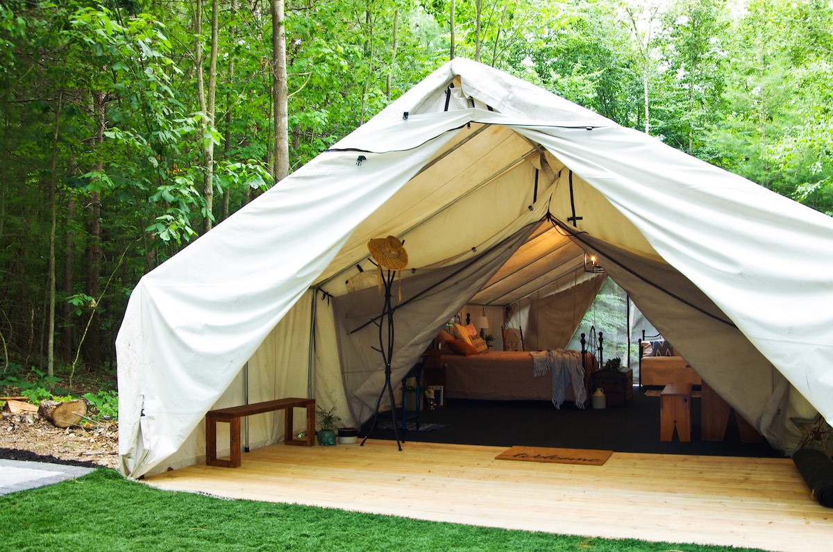 A spacious glamping tent is shown, nestled among trees. Wooden flooring is visible at the entrance, leading to a well-arranged interior featuring a bed and rustic furnishings. Natural light filters through the fabric, creating a serene ambiance for guests to enjoy the outdoors.