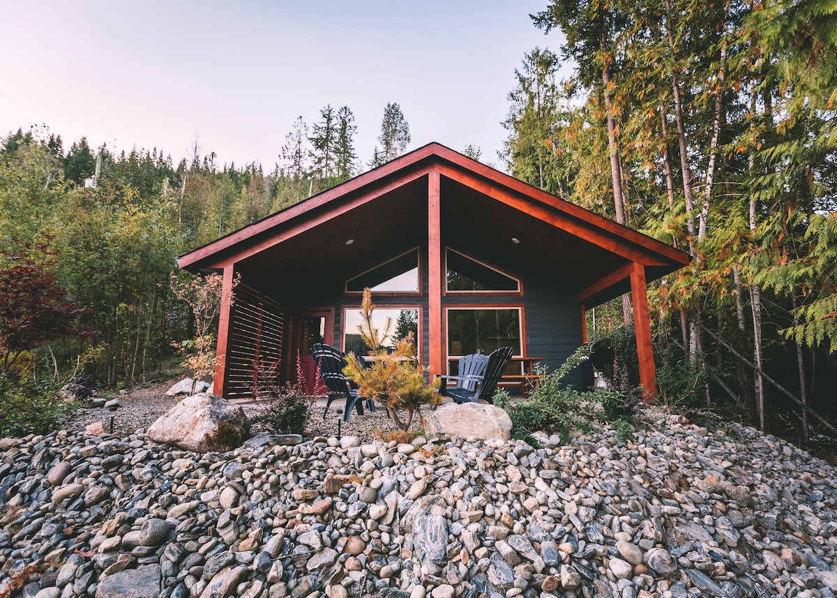 A modern cabin is nestled among trees and rocks, featuring a triangular roof and large windows. Adirondack chairs are arranged outside, surrounded by natural stone landscaping. The cabin's exterior showcases a dark color with bright red accents, blending harmoniously with the tranquil forest setting.
