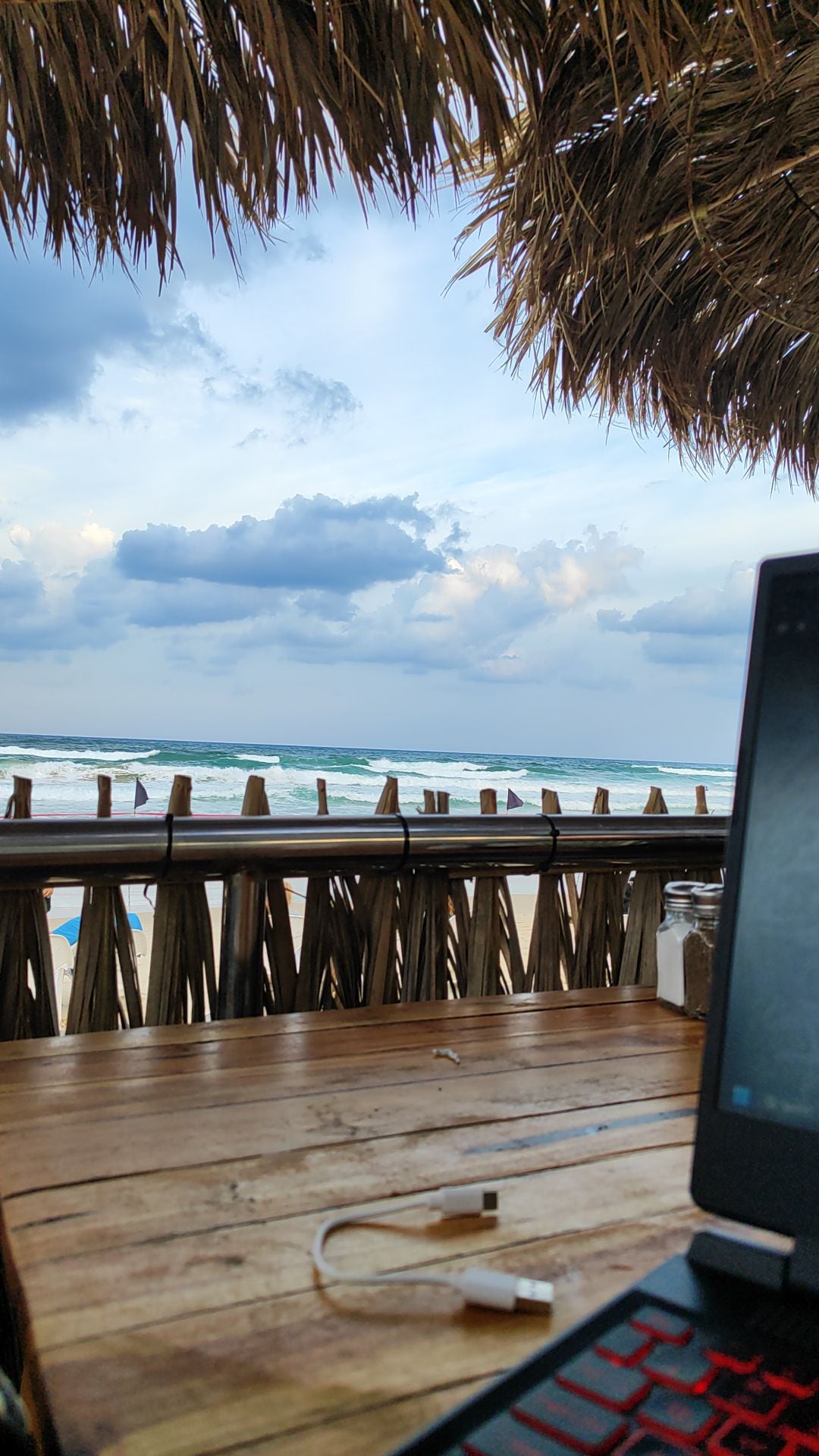 A wooden table is shown with a laptop and a charger, positioned under a thatched roof. In the background, the sea is visible with gentle waves, under a cloudy sky. The beach is partially seen, enhancing a sense of proximity to the ocean.
