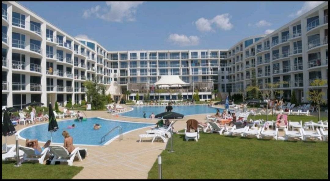 A large outdoor area highlights two swimming pools surrounded by sun loungers and umbrellas. Lush green lawns provide space for relaxation. The modern architecture of the surrounding building features multiple balconies, creating an inviting atmosphere within the resort complex.