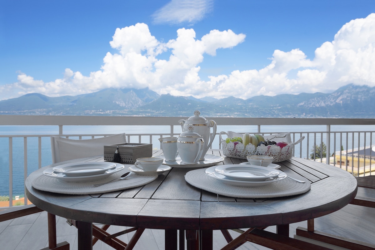 A wooden table is set for breakfast with white dishware and a basket of colorful fruits. The view beyond overlooks Lake Garda, framed by distant mountains and a bright blue sky with scattered clouds.