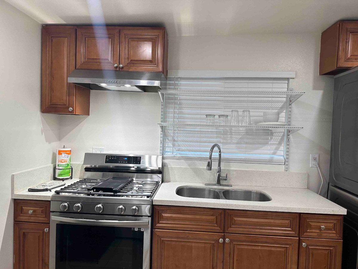 A modern kitchen is depicted with wooden cabinets and granite countertops. A stainless steel gas stove and range hood are seen, complemented by a sink with a sleek faucet. Open shelving displays glassware, illuminated by natural light through the window.