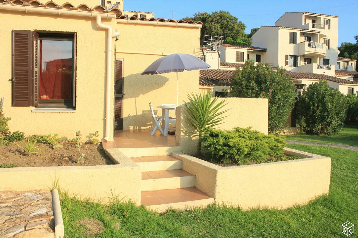 A small patio area features a white table and two chairs under a blue umbrella. Steps lead up to the entrance, surrounded by greenery and a low stone wall. The light yellow exterior of the building contrasts with the lush grass and nearby structures.