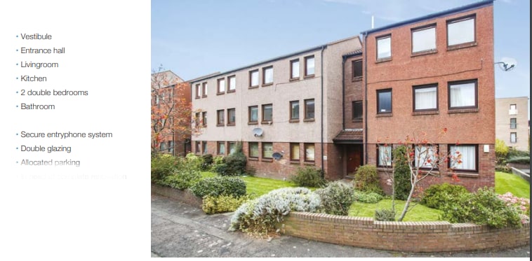 The exterior of the first-floor flat is shown, featuring a brick facade with several windows. Greenery lines the pathway leading to the entrance, and an allocated parking space is visible in the nearby area. The overall setting is quiet with a residential ambiance.