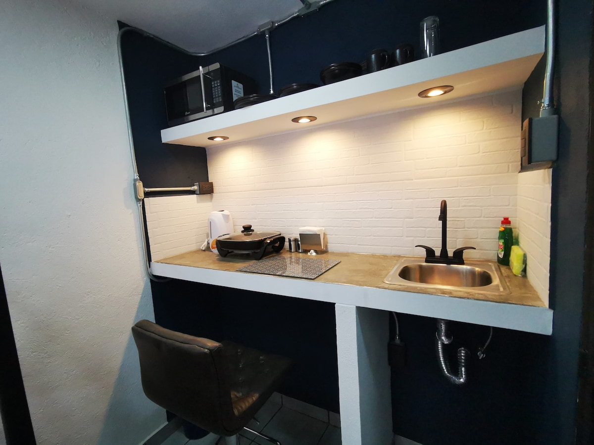 A compact kitchenette is equipped with essential appliances, including a microwave and stovetop. A sink is positioned beneath a shelf illuminated by warm lights. Dark cabinetry contrasts against the white brick backsplash, while a stool is placed at the counter for seating.