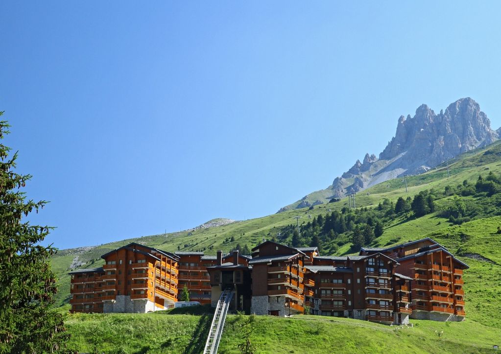 The residence 'Le Tueda' is situated in a verdant landscape, framed by a mountain backdrop. The structure features a combination of wood and stone design, with multiple balconies on the upper floors, offering an inviting appearance that blends with the natural surroundings.