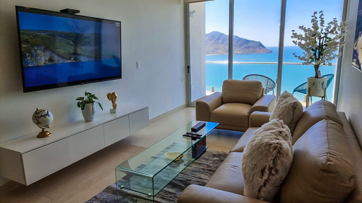 A bright living area is presented with a large flat-screen TV mounted on a white wall. A glass coffee table sits on a textured rug in front of a spacious beige sofa, while a scenic view of the ocean and mountains is framed by large glass windows.