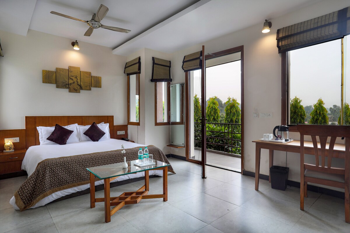 A well-appointed room is seen, featuring a spacious bed with a decorative brown throw. Large windows allow natural light to flow in, revealing a private balcony with greenery views. A wooden table is positioned adjacent to the bed, accompanied by a kettle and glassware.