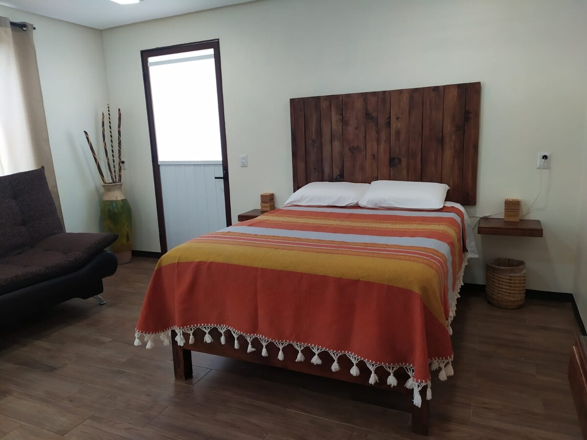 A spacious room features a wooden headboard and a colorful striped blanket draped over the bed. A sofa bed is positioned nearby, and wooden side tables with decorative elements are present. Natural light filters in through a window, complemented by earthy tones in décor.