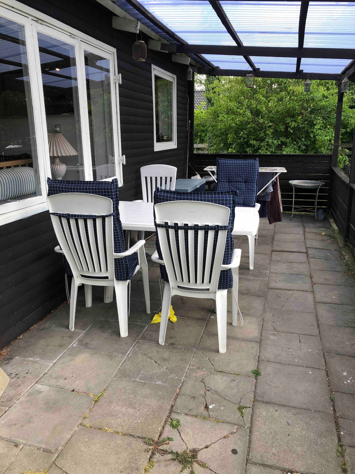 An outdoor patio area features a white dining table accompanied by six plastic chairs with blue cushions. A retractable roof provides shade, while potted greenery offers a natural backdrop. Nearby, a clothesline and additional seating are included, enhancing the space's functionality.