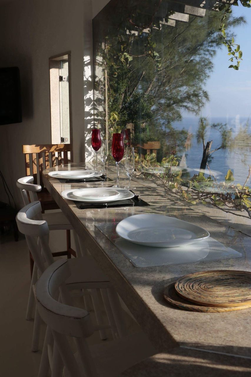 A dining area features a long counter with several white plates and elegant glassware, set against a backdrop of large windows offering views of the beach. Sunlight streams in, reflecting off the polished surfaces, while greenery is visible outside.