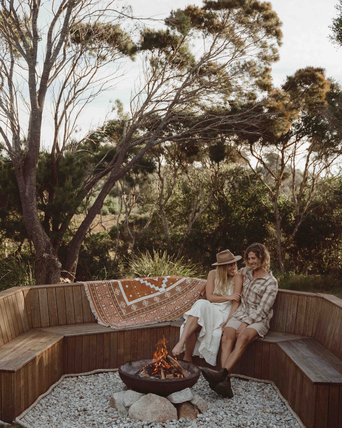 A circular wooden seating area surrounds a central fire pit, situated among native trees and greenery. A decorative rug adds color to the space, which is designed for relaxation and conversation. The natural surroundings contribute to a serene and private atmosphere.