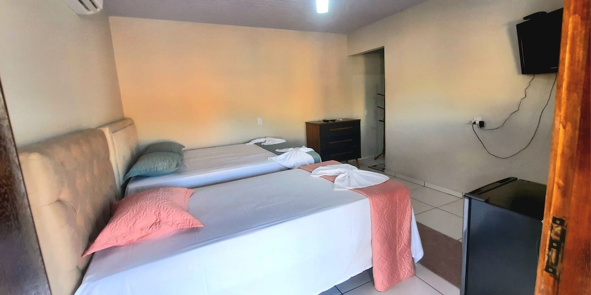 The sleeping area features two spacious beds, one adorned with a light pink accent pillow. A dresser and a small refrigerator are present, along with a wall-mounted television for entertainment. Natural light filters in from a window, creating a comfortable environment.