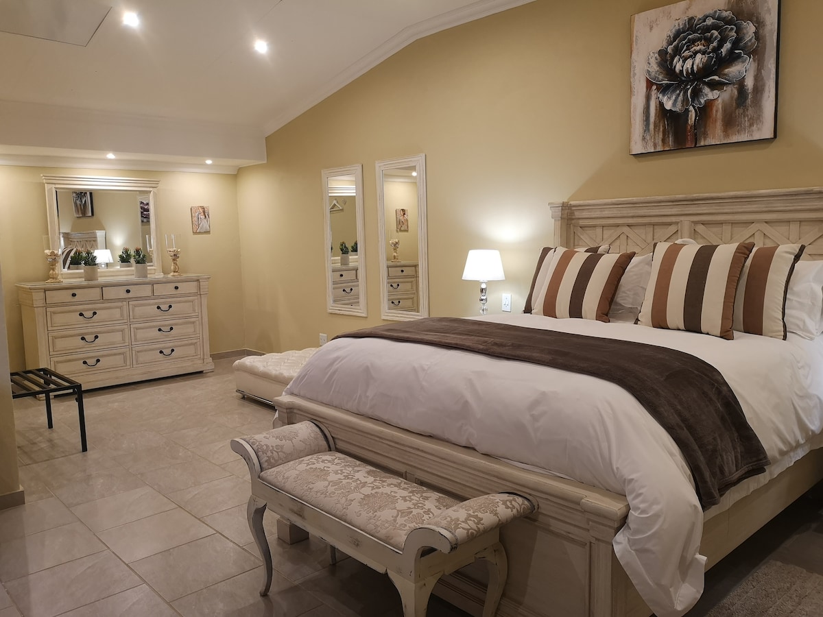 A spacious room features a beautifully crafted bed with decorative pillows, accompanied by a matching bench at the foot. A dresser and mirrors line one wall, contributing to the room's elegant decor, while soft lighting creates a calm and inviting atmosphere.