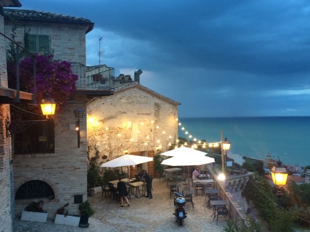 A charming outdoor terrace is visible, featuring several tables with umbrellas. Warm lights hang above, creating a cozy ambiance. The backdrop includes a coastline with blue waters under a slightly overcast sky, and stone buildings adorned with greenery.