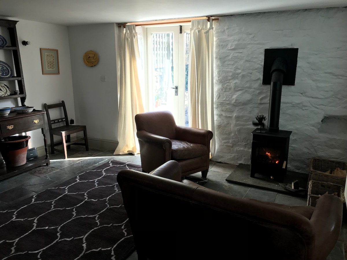 A cozy lounge space is visible, featuring two armchairs and a wood burning stove, which provides warmth to the room. Light flows in through the glass door, leading to the garden. Antique furnishings, including a wooden dresser, are positioned along the stone wall.