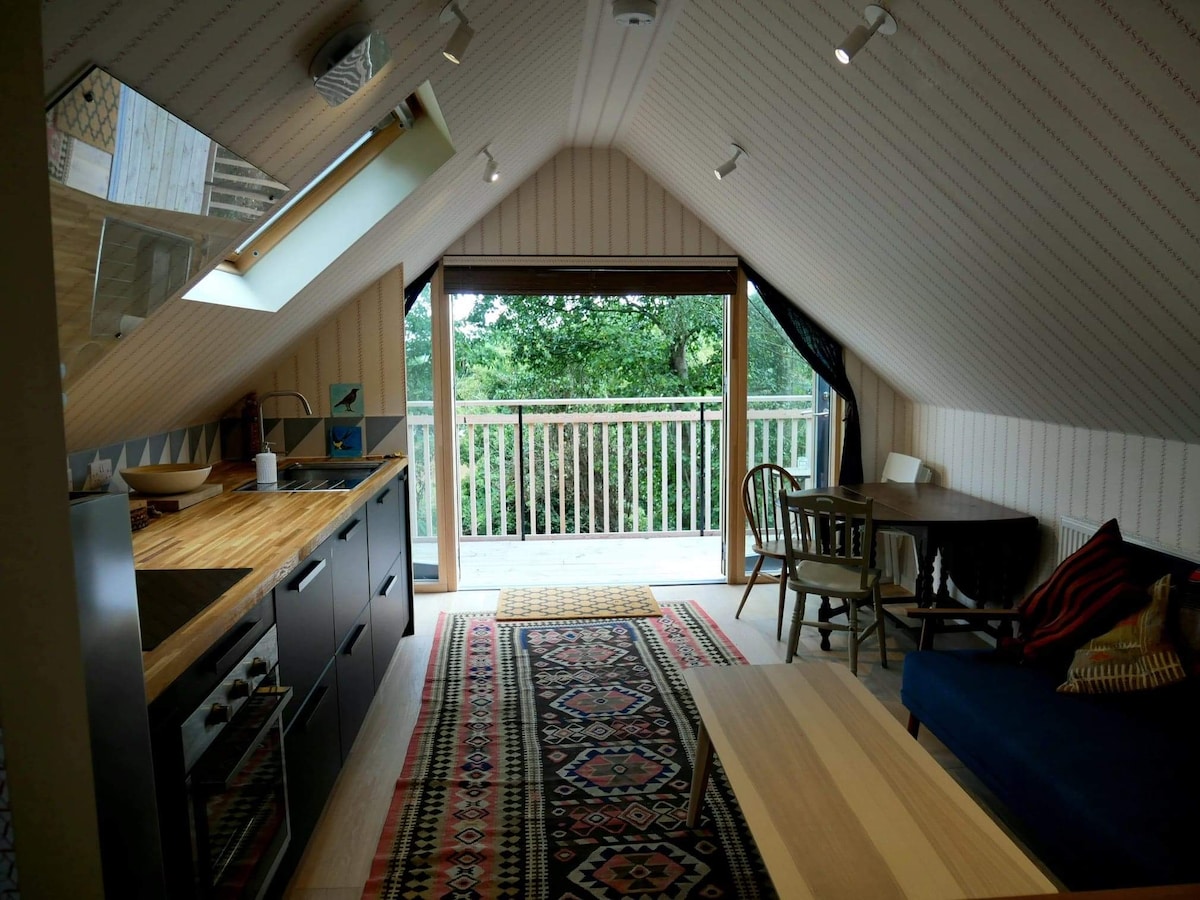 The studio apartment features a modern kitchen with a wooden countertop and sleek black cabinetry. A cozy seating area includes a dark sofa and a small dining table by the expansive window, offering views of the surrounding greenery. Natural light fills the space through angled skylights.