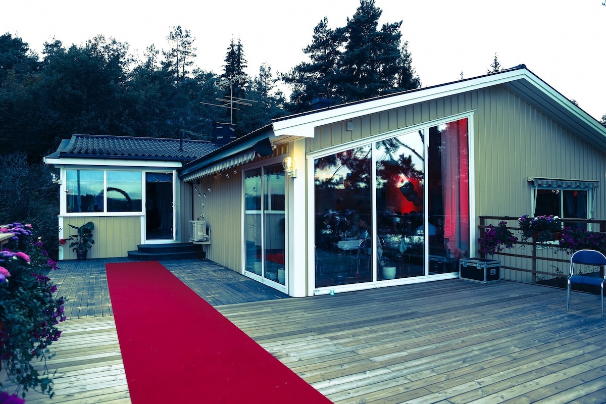 An exterior view of the main house is displayed, showing large glass windows and a wooden terrace. A red carpet leads to the entrance, surrounded by flowering plants. The setting is framed by trees in the background, enhancing the natural surroundings.
