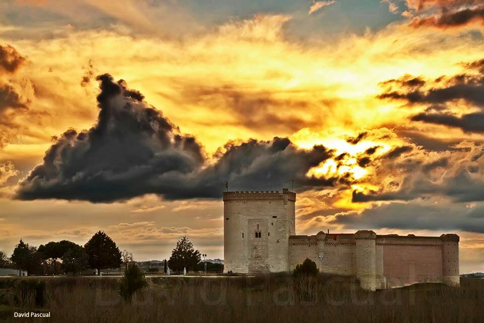 A historical castle stands against a dramatic sky, featuring a mix of orange, yellow, and gray clouds at sunset. The structure is characterized by its sturdy stone walls and towers, surrounded by sparse vegetation and an open field.