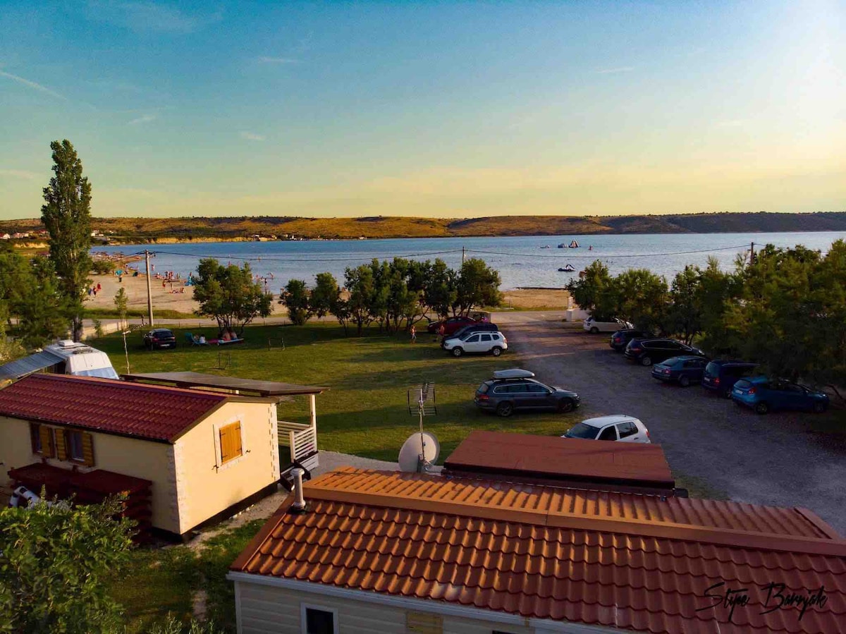 A view from above captures the mobile house with a red-tiled roof and surrounding greenery. A sandy beach and calm water are visible in the distance, with scattered cars parked nearby. The scene conveys a relaxed outdoor atmosphere perfect for family outings.