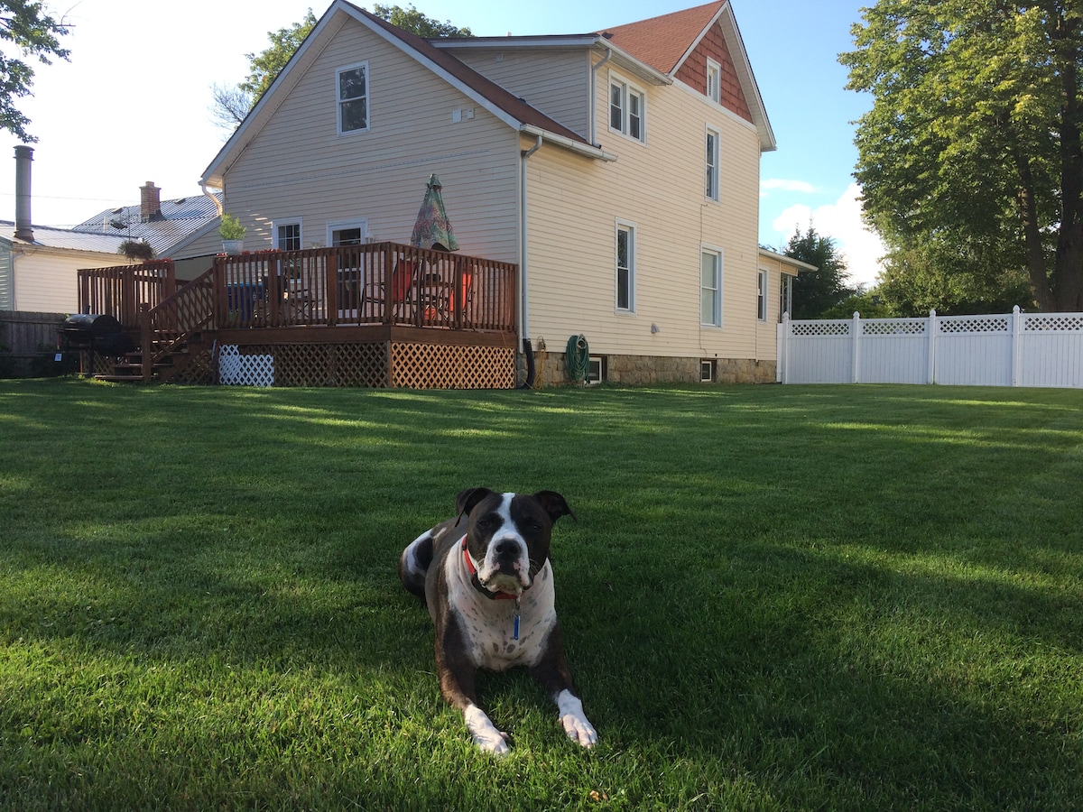 A spacious backyard is featured, with a well-maintained lawn and a friendly dog lounging on the grass. The two-story house is visible in the background, showcasing a wooden deck and surrounded by trees. The property is enclosed by a white picket fence.