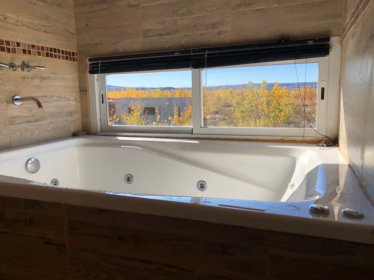 A spacious hot tub is positioned against a large window, offering a view of the surrounding landscape. The tub features multiple jets, while the natural light enhances the warm tones of the wooden wall finishes and the decorative strip above the tub.