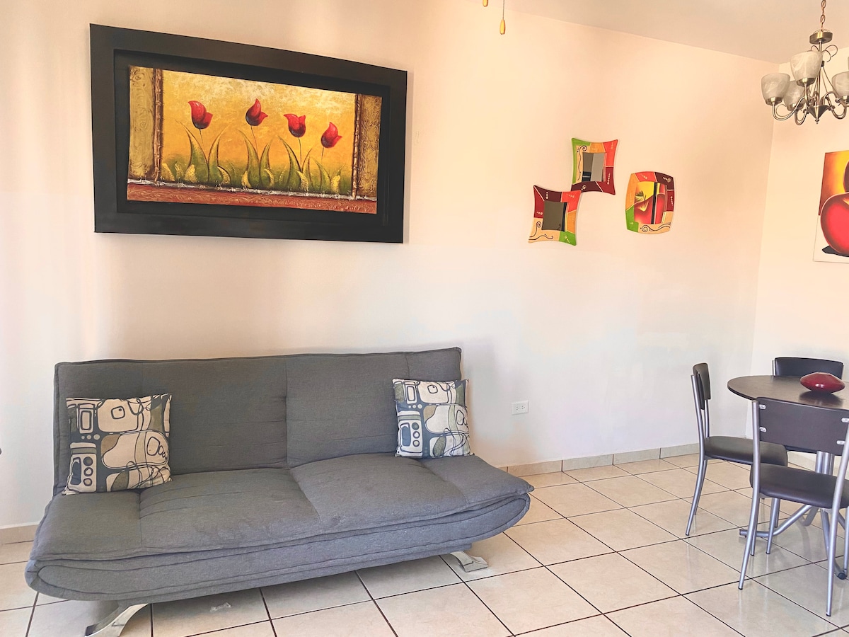A gray sofa bed is positioned against a light-colored wall, adorned with vibrant wall art featuring flowers and abstract designs. A small circular dining table with four metal chairs is visible in the corner, creating a functional and inviting living space.