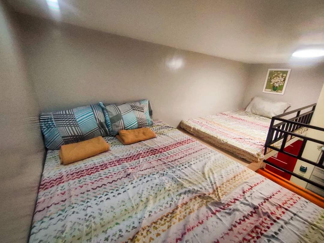 Two spacious beds are arranged in a loft-style layout, each adorned with patterned bedding in soft hues. Pillows and folded towels are placed on the beds, providing a welcoming touch. Ambient lighting enhances the room's cozy atmosphere.