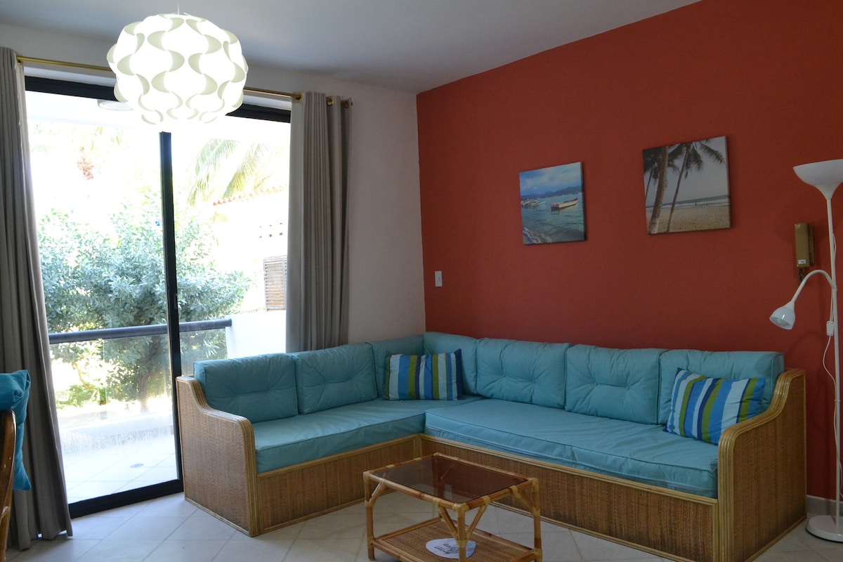 A spacious living area features a comfortable, L-shaped turquoise sofa adorned with colorful cushions. Large glass doors allow natural light to fill the room, providing a view of the outdoors. Light-colored curtains frame the windows, and a modern pendant light hangs from the ceiling.
