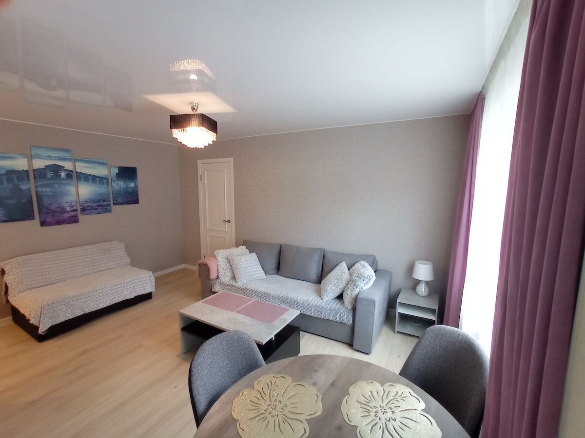 A spacious living area is highlighted by a neutral color palette, featuring a gray sofa and a daybed. A round dining table with soft chairs sits nearby, while large windows provide ample natural light. Decorative artwork adorns the wall, adding a touch of color.