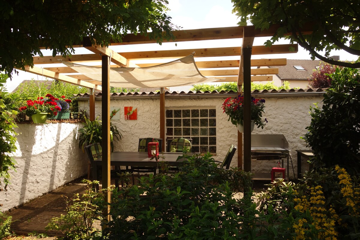 The garden area features a wooden pergola covered with a light shade, providing a space for outdoor dining. A table and several chairs are set amidst lush greenery and colorful flower pots, with a grill positioned nearby for barbecue evenings.