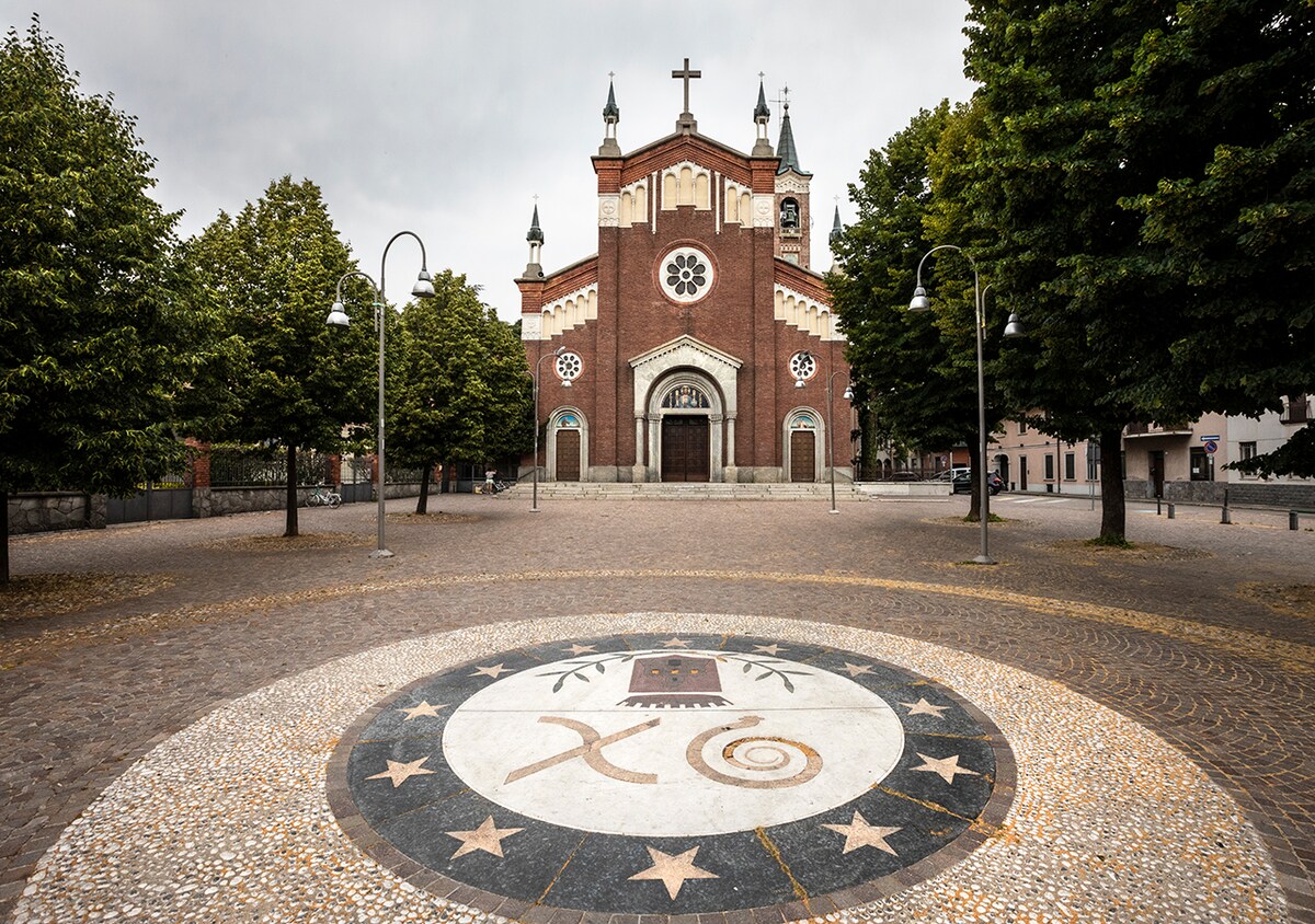 A historic church, characterized by red brick architecture, stands prominently. Circular patterns pave the foreground, featuring designs and symbols. Lush green trees line the plaza, with street lamps illuminating the area, providing a serene setting for visitors.