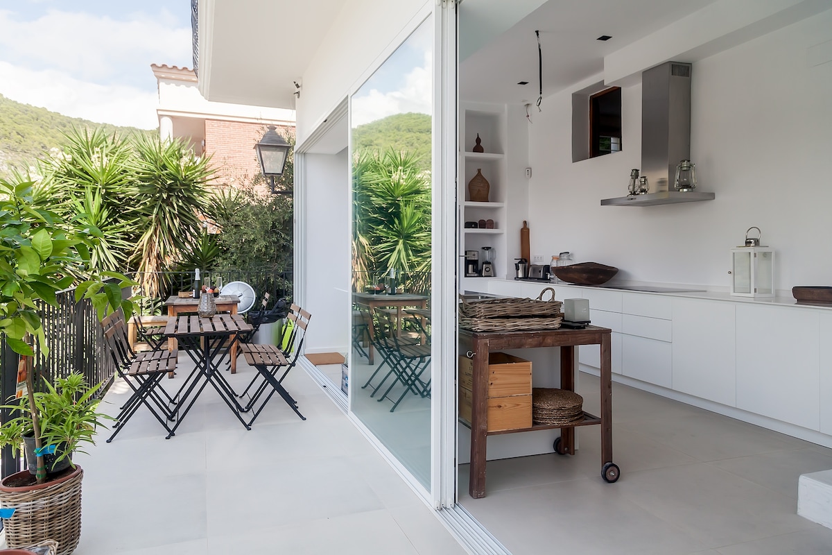 An open outdoor space is showcased, featuring a wooden table surrounded by matching folding chairs. Lush greenery provides a tranquil backdrop, while a small cart adds functionality. Large glass doors offer a view into a modern kitchen area, creating a seamless connection between indoor and outdoor spaces.