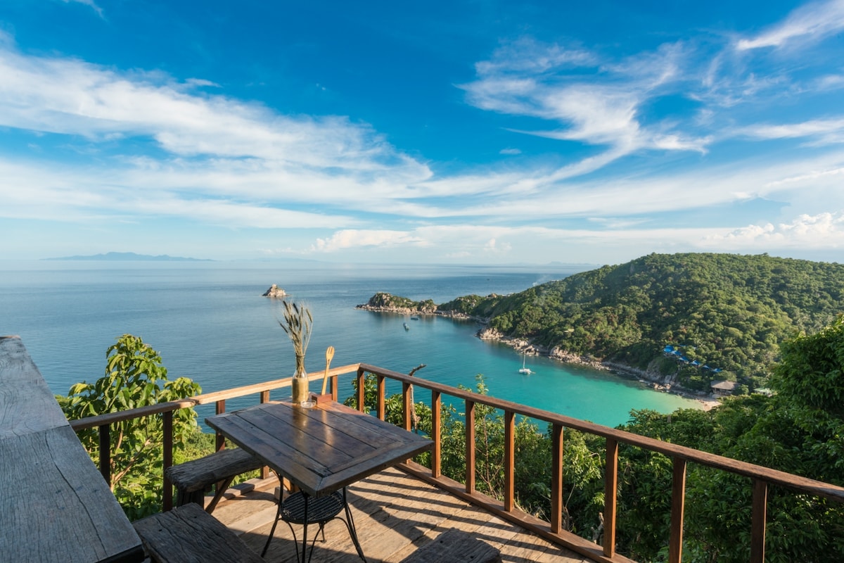 A wooden terrace overlooks the calm turquoise waters and the lush coastline of Koh Tao. The scene is enhanced by a clear blue sky with scattered clouds, while a rustic table and chairs provide a picturesque spot to enjoy the view.
