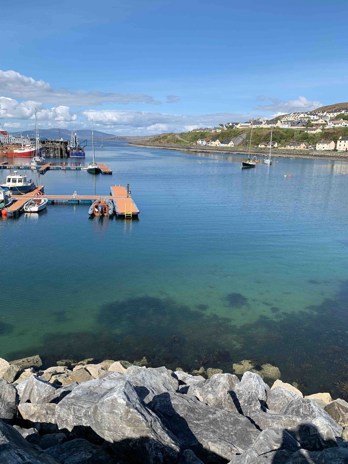 A serene view of Mallaig harbour is presented, showcasing calm waters reflecting the sky. A wooden dock extends into the water, with several boats moored nearby. Surrounding hills and the village buildings are visible in the distance, framed by a clear blue sky with scattered clouds.