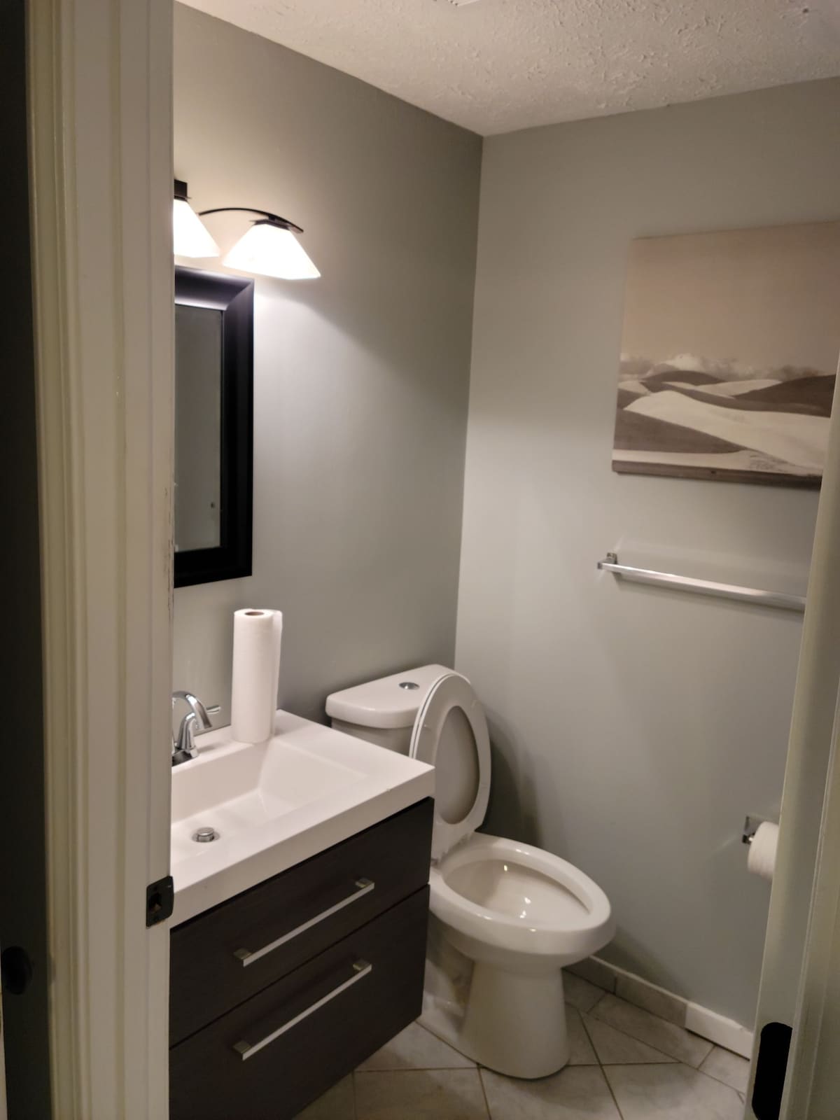 A compact bathroom is presented, featuring a modern sink with a vanity and two drawers below. A toilet is positioned beside the sink. A framed artwork adorns the wall, while a paper towel roll is available on the countertop. Soft lighting is provided by wall fixtures.