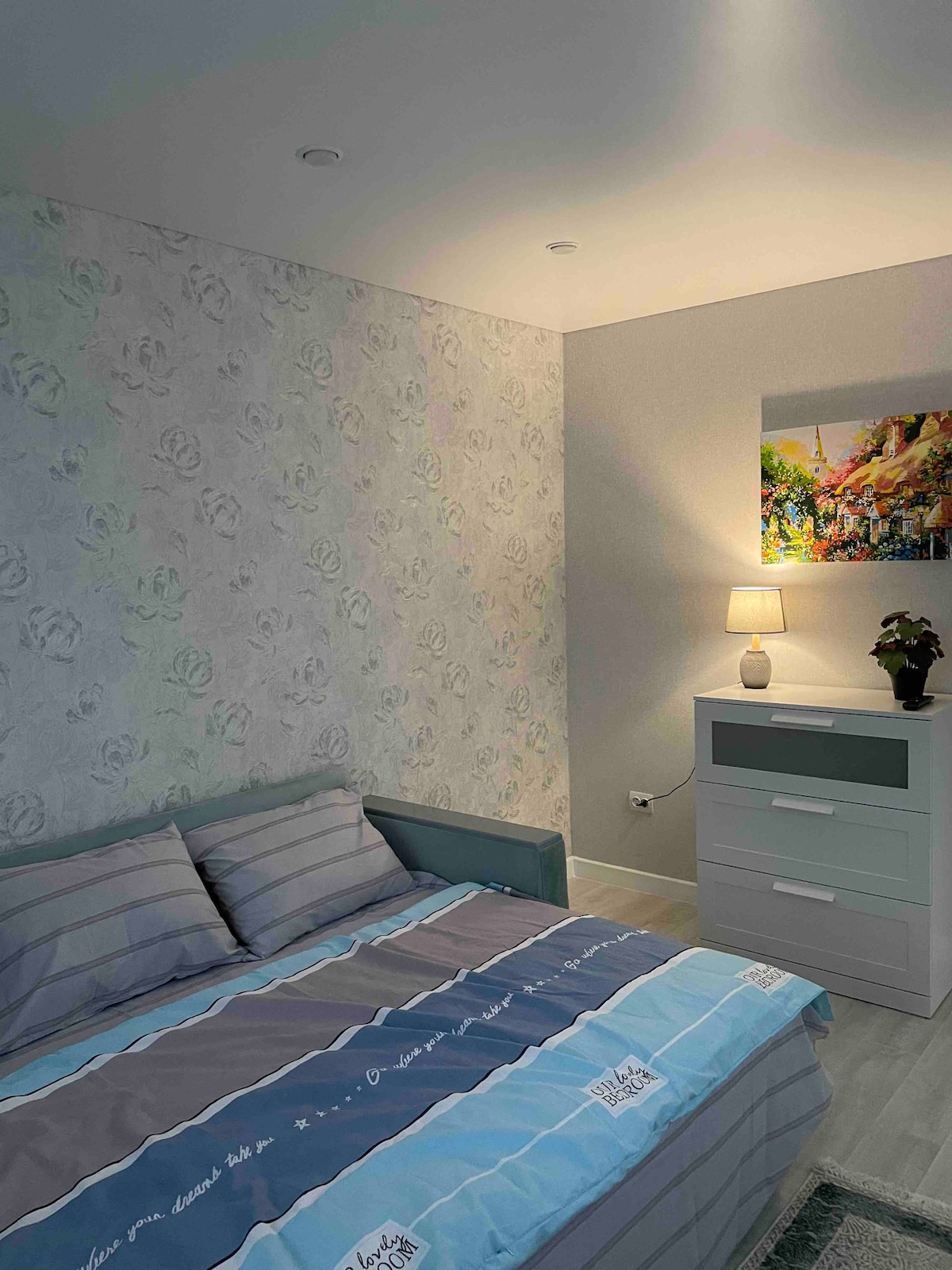 A well-lit bedroom features a spacious bed with striped bedding in shades of blue and gray. A small bedside table with a lamp and a decorative plant sits next to the bed. The wall is adorned with a floral wallpaper and a colorful artwork above the dresser.