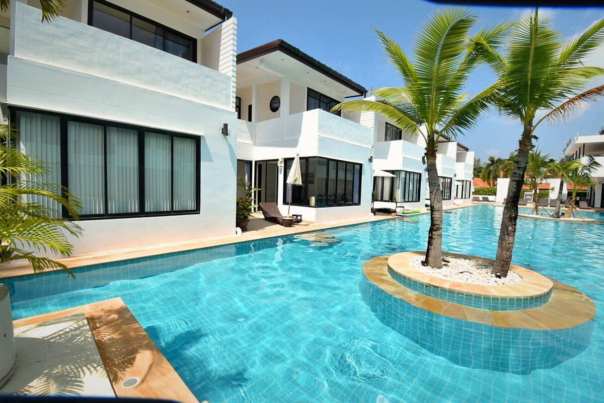 A vibrant swimming pool with crystal-clear water is framed by palm trees and modern townhouses. The pool features a circular island with decorative stones and is surrounded by spacious patios, providing an inviting atmosphere for relaxation.