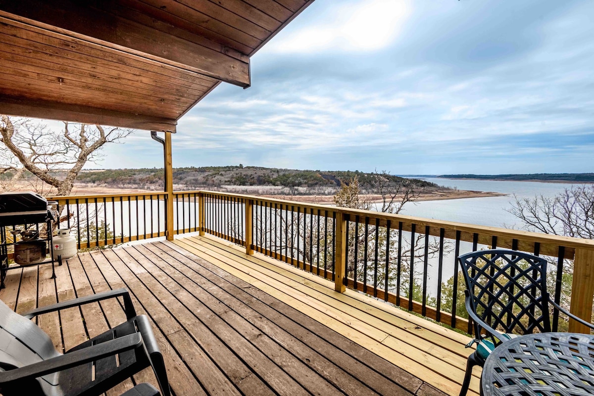 A spacious deck overlooks Baldwin Cove on Tuttle Creek Lake, with wooden flooring and railings. Seating arrangements include two chairs and a round table, providing an inviting space to enjoy the serene views of the surrounding landscape and tranquil water.