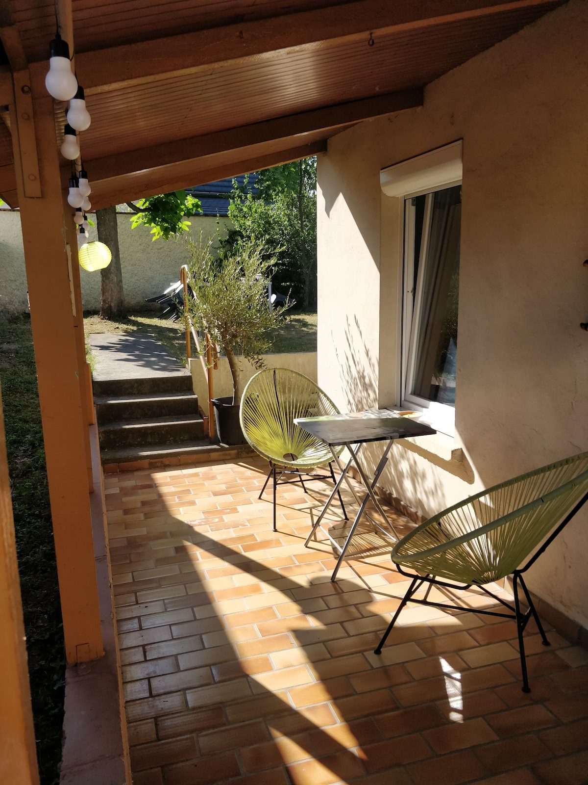 An outdoor seating area is presented with two green chairs and a small table under a wooden overhang. Natural light streams across the tiled patio, highlighting the surrounding greenery and steps leading to the garden.