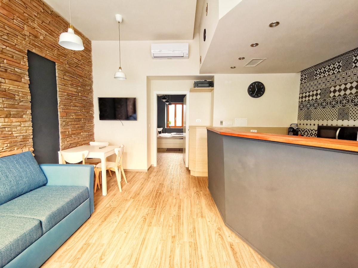 A modern interior features a cozy blue sofa and a light wooden dining table with chairs. Wall textures include natural stone and patterned tiles. An air conditioning unit and a clock are visible, with a view leading to a bedroom area in the background.