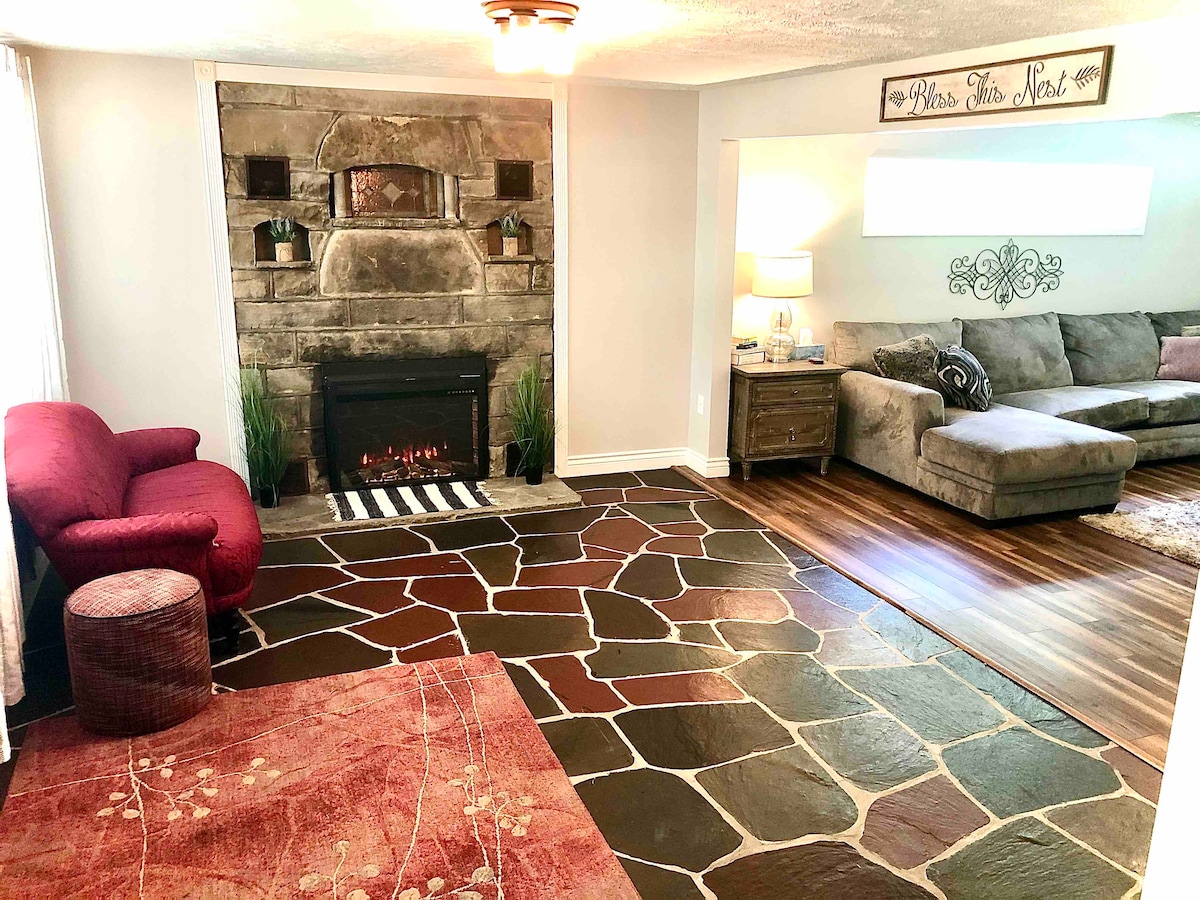 The living room features a stone fireplace as a focal point, surrounded by a modern sectional couch and a red accent chair. Natural light enters through large windows, highlighting the unique stone flooring and warm wood tones.