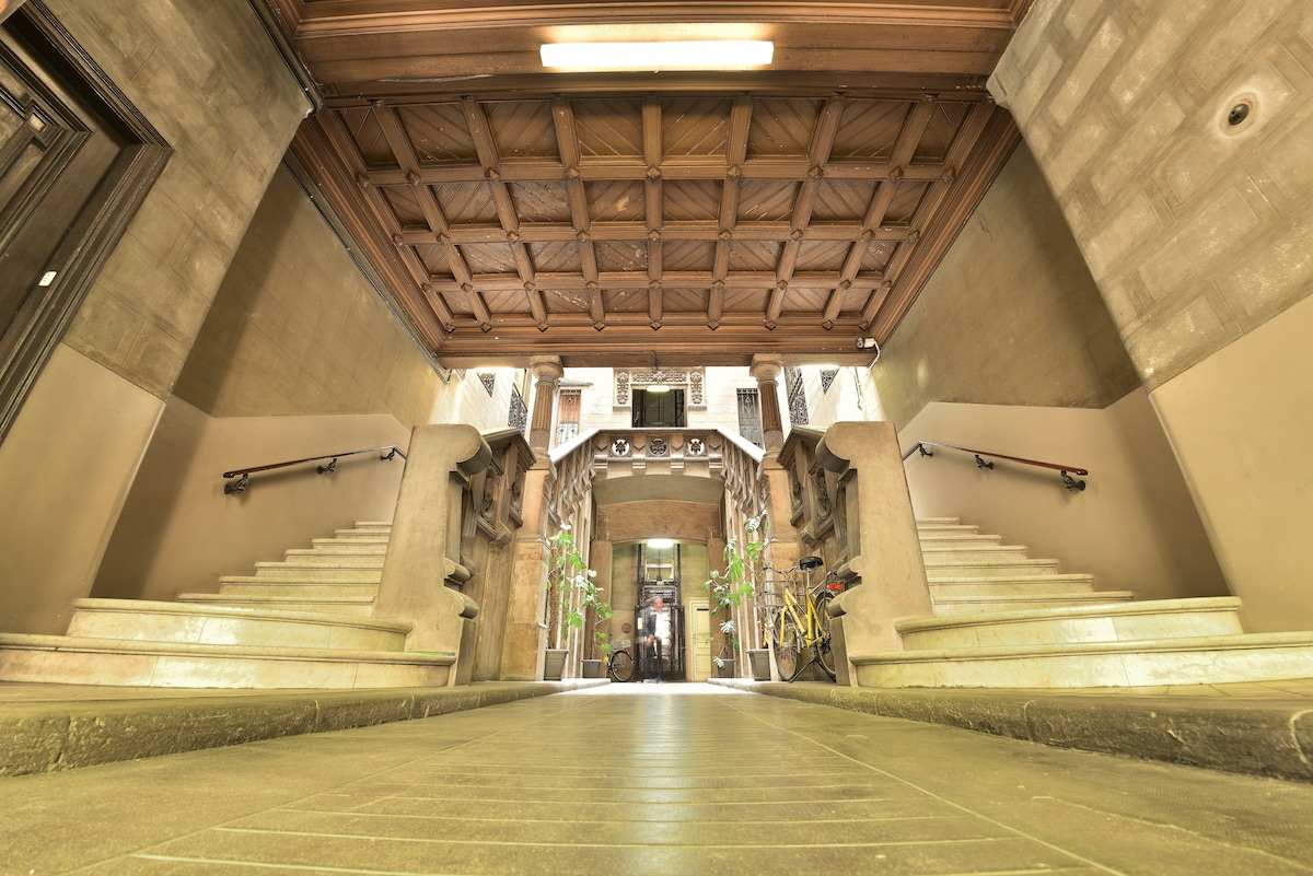 The entrance hallway features a high wooden coffered ceiling with intricate details. Stone stairs lead up on both sides, flanked by decorative columns and greenery. An inviting atmosphere is created by soft lighting illuminating the pathway leading into the building.