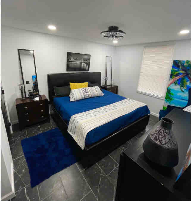 A well-appointed bedroom showcases a spacious bed with dark bedding and decorative pillows. Soft lighting enhances the room's atmosphere, while accent furniture includes a bedside table and a tall mirror. A plush rug adds comfort beneath the bed, and natural light filters through a partially covered window.