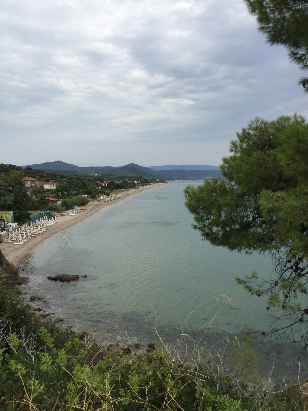 A tranquil beach scene is depicted, featuring a sandy shoreline bordered by greenery. The calm sea reflects a muted sky, while distant hills provide a scenic backdrop. Sun loungers are visible along the beach, suggesting a welcoming space for relaxation.
